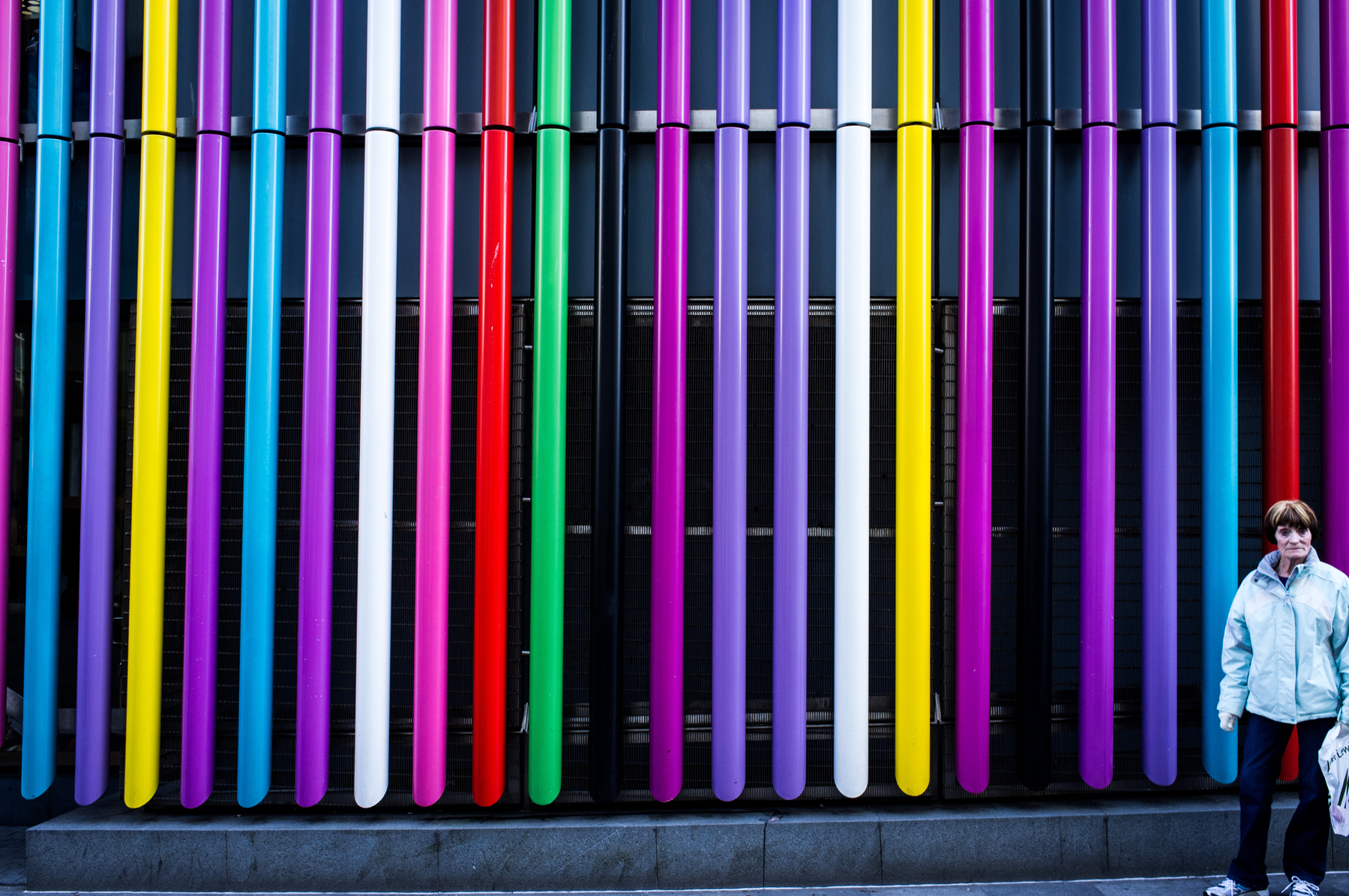 Auto-generated description: A colorful wall with vertical bars in various shades is accompanied by a woman standing on the right.