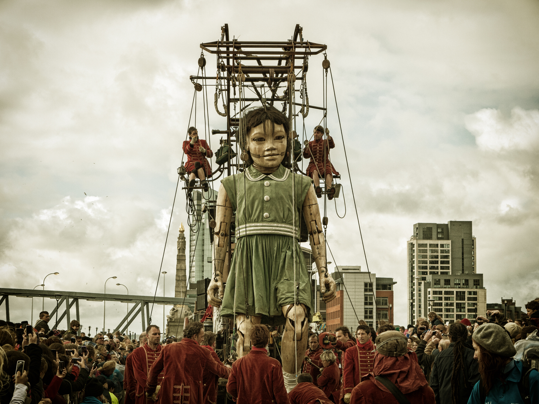 Auto-generated description: A large, lifelike puppet of a girl is being maneuvered by operators in red clothing amidst a crowd in an urban setting.